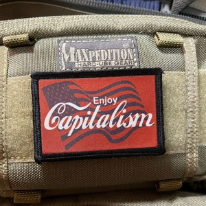 May include: A red, white, and blue American flag patch with the text "Enjoy Capitalism" in white lettering. The patch is attached to a green tactical backpack with a black Maxpedition Hard-Use Gear label.