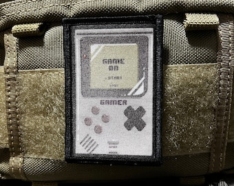 Puede incluir: Un parche blanco y negro con una imagen pixelada de una consola de videojuegos portátil. La pantalla muestra el texto "GAME ON" y la consola tiene un botón etiquetado como "START". La consola está etiquetada como "GAMER".