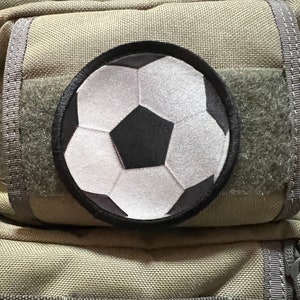 Op de afbeelding: Een close-up van een beige Maxpedition Hard-Use Gear rugzak met een zwart-witte voetbalpatch op de voorkant.