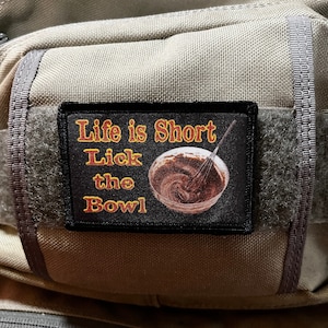 May include: A black and white patch with the text "Life is Short Lick the Bowl" in orange and yellow. The patch features a bowl of chocolate batter with a whisk in it.