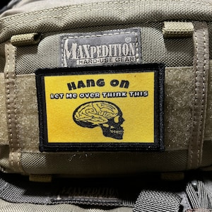 May include: A black and yellow patch with the text "HANG ON LET ME OVER THINK THIS" and a skull with a brain inside. The patch is attached to a green Maxpedition bag.