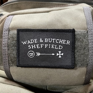 Op de afbeelding: Een zwarte patch met witte tekst die "Wade & Butcher Sheffield" luidt met een cirkel met de letter "B" en een pijl die naar rechts wijst. Een wit kruis bevindt zich rechts van de pijl.