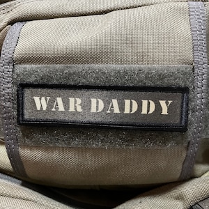 Könnte beinhalten: Ein grauer taktischer Rucksack mit einem schwarzen Patch, auf dem "WAR DADDY" steht.