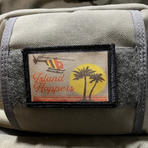 May include: A beige fabric patch with black stitching and a black border. The patch features a cartoon helicopter with orange and black stripes, palm trees, and the text "Island Hoppers".