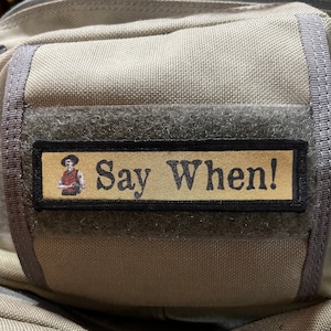 Könnte beinhalten: Ein beigefarbener Stoffrucksack mit einem schwarzen und goldenen Patch, auf dem "Say When!" steht und ein Cartoonbild einer Person mit Cowboyhut zu sehen ist.