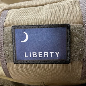 May include: A tan fabric backpack with a black patch featuring a blue crescent moon and the word "LIBERTY" in white.