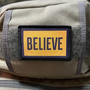 May include: A beige tactical bag with a black and yellow patch that says "BELIEVE" in purple lettering.