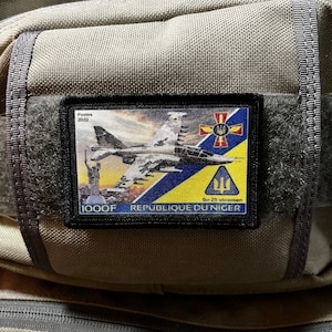 May include: A black and yellow patch with a blue and yellow Ukrainian flag, a white cross, and a gray fighter jet. The patch also has the text "1000F Republique du Niger" and "Su-25 ukrainian".