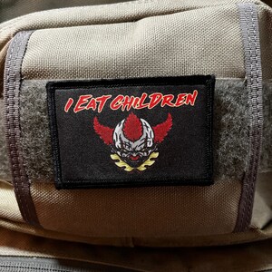 May include: A black patch with white stitching and the text "I EAT CHILDREN" in red letters. A red and white clown face with yellow wings is in the center of the patch.