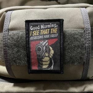 May include: A black and yellow patch with the text "Good Morning, I See That The Assassins Have Failed" and a cartoon image of a hand holding a gun.