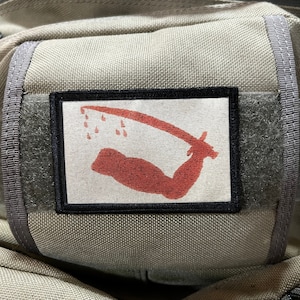 May include: A gray backpack with a black and white patch featuring a red stylized arm with a sword and dripping blood. The patch is sewn onto the front of the backpack.