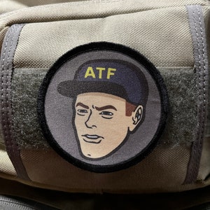May include: A gray and black circular patch with a cartoon illustration of a person wearing a blue baseball cap with the text "ATF" in yellow letters. The person has brown hair and a serious expression.