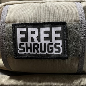 May include: A beige backpack with a black patch that says "FREE SHRUGS" in white letters.