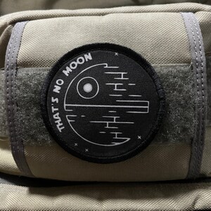 May include: A black and white patch with the text "That's no moon" and a stylized image of the Death Star from Star Wars. The patch is attached to a beige backpack.