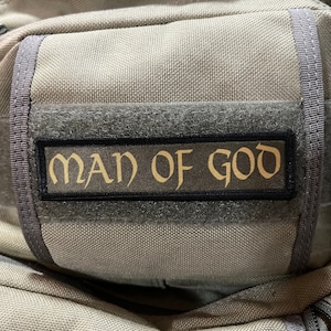 Könnte beinhalten: Ein grauer taktischer Rucksack mit einem schwarzen Patch, auf dem in goldenen Buchstaben "MAN OF GOD" steht.