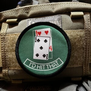 May include: A green and black embroidered patch with a playing card design. The cards show a 5 of clubs, a 6 of hearts, and a 9 of spades. The text on the patch reads "I'd Hit That".