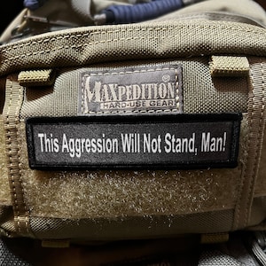 May include: A close-up of a green Maxpedition bag with a black patch that reads "This Aggression Will Not Stand, Man!" The bag has a black Maxpedition logo with the text "Hard-Use Gear".
