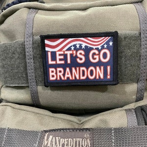May include: A green Maxpedition backpack with a black patch that says "Let's Go Brandon!" with an American flag design. The backpack has a black strap and a black label that says "Maxpedition Hard-Use Gear".