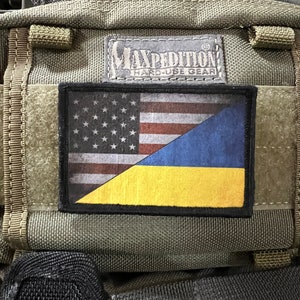 May include: A green tactical backpack with a black patch featuring the American and Ukrainian flags. The patch has the text "Maxpedition Hard Use Gear" above the flags.
