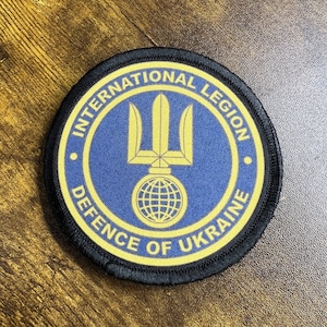May include: A black and yellow round patch with the text "International Legion Defence of Ukraine" and a blue background with a gold trident and globe.