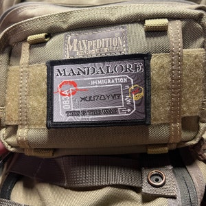 May include: A green Maxpedition hard-use gear backpack with a black patch that says "MANDALORE" with the text "IMMIGRATION" and "THIS IS THE WAY" on it.