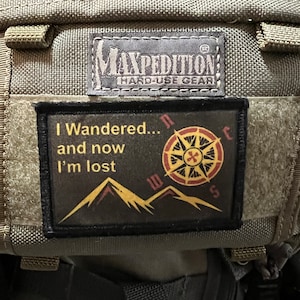 Puede incluir: Una mochila verde de estilo militar con un parche negro que dice "I Wandered... and now I'm lost" con una brújula y montañas. La mochila tiene una etiqueta negra que dice "Maxpedition Hard-Use Gear".