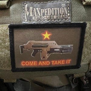 以下が含まれることがあります： 赤い星と黒と銀のライフルが描かれた黒いパッチで、オレンジ色の「COME AND TAKE IT」という文字が書かれています。