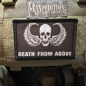 May include: A black and white patch with a skull and wings. The patch is sewn onto a green tactical bag. The patch says "Maxpedition Hard-Use Gear" and "Death From Above".