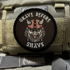 May include: A black and white patch with a cartoon viking wearing a horned helmet and a beard. The patch says "Grave Before Shave" in white text.