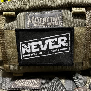 May include: A green and black tactical bag with a black patch that says "NEVER TELL ME THE ODDS" and a black label that says "MAXPEDITION HARD-USE GEAR".