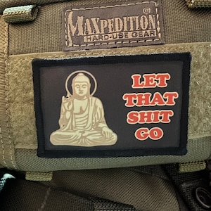 Pode incluir: Uma bolsa verde de estilo militar com um remendo preto que diz "Let That Shit Go" e apresenta uma imagem de desenho animado de um Buda meditando. A bolsa tem uma etiqueta Maxpedition Hard-Use Gear.