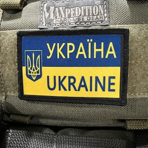 May include: A green military-style bag with a black patch that reads "УКРАЇНА UKRAINE" in yellow and blue lettering. The bag has a black label that reads "MAXPEDITION HARD-USE GEAR".