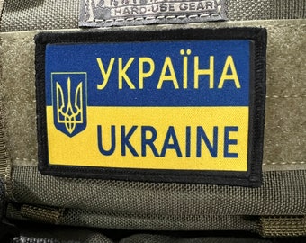 Puede incluir: Una bolsa verde de estilo militar con un parche negro que dice "УКРАЇНА UKRAINE" en letras amarillas y azules. La bolsa tiene una etiqueta negra que dice "MAXPEDITION HARD-USE GEAR".