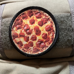 Könnte beinhalten: Ein runder, schwarzer Patch mit einem Design einer Pepperoni-Pizza. Die Pizza hat einen gelben Käsegrund und rote Pepperoni-Scheiben.