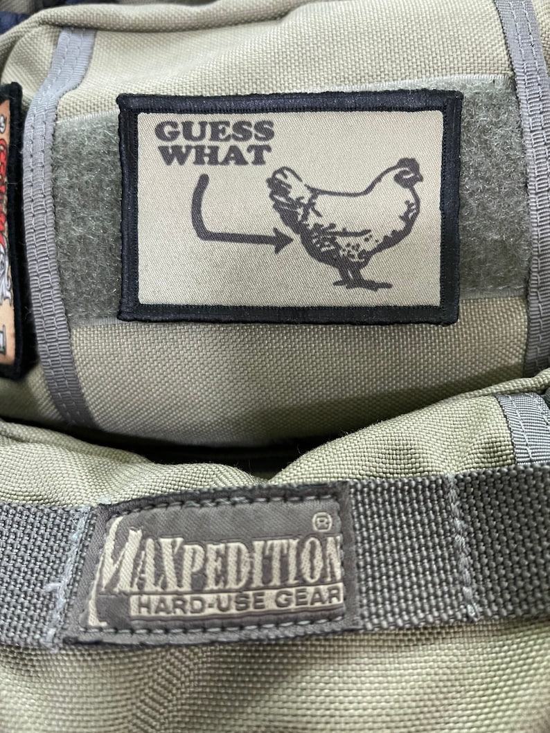 Pode incluir: Uma bolsa de lona verde com um remendo preto que diz "GUESS WHAT" e apresenta uma galinha com uma flecha apontando para sua traseira. A bolsa tamb&eacute;m tem um remendo preto que diz "MAXPEDITION HARD-USE GEAR".