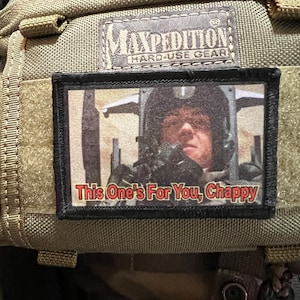 May include: A black and green patch with a picture of a person in a helmet and the text "This One's For You, Chappy". The patch is attached to a green Maxpedition backpack.