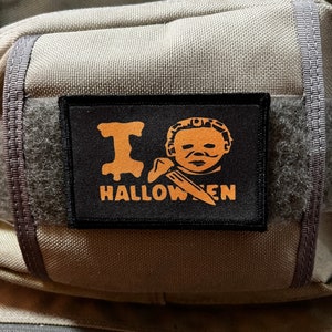 May include: A black patch with orange text that reads "I  HALLOWEEN" and a graphic of a person with a mask. The patch is attached to a gray fabric bag.