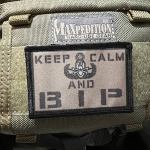 Puede incluir: Una mochila Maxpedition Hard-Use Gear de color beige y negro con un parche negro que dice "Keep Calm and 13 112" con una insignia militar en el centro.