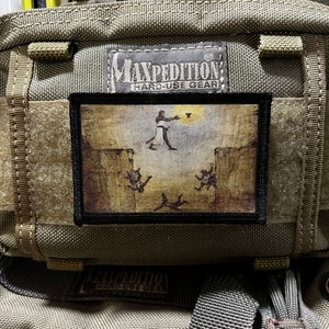 May include: A Maxpedition Hard-Use Gear patch with a brown background and a black border. The patch features a black and white illustration of a figure in a robe holding a staff, with three figures falling from the sky. The text "Maxpedition Hard-Use Gear" is printed in white on a black background.