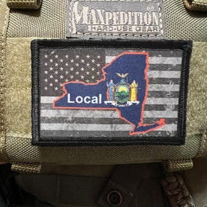 May include: A black and gray patch with a faded American flag design and an outline of the state of New York. The patch has the word "Local" printed in white text on the state outline. The patch is attached to a green tactical bag with the text "EXPEDITION HARD-USE GEAR" printed on it.