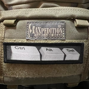 May include: A tan Maxpedition Hard-Use Gear backpack with a black patch that says "Ctrl, Alt, Del" in white text.