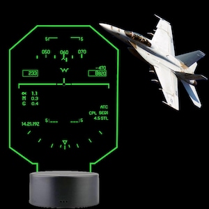 May include: A decorative lamp featuring a glowing green acrylic panel etched with a fighter jet heads-up display interface, including numerical data and flight symbols, mounted on a solid black circular base against a dark background.