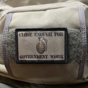 Puede incluir: Un parche negro con texto blanco que dice "Close Enough For Government Work". Un dibujo animado de una granada está en el centro del parche.