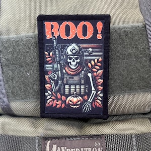 Puede incluir: Un parche negro con un soldado esqueleto sosteniendo un rifle y una calabaza. El parche tiene el texto "BOO!" en letras rojas.