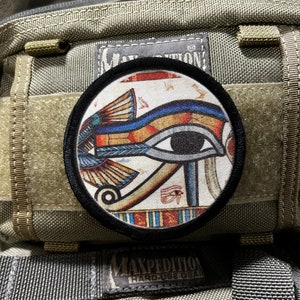 May include: A black and white patch featuring a colourful image of the Eye of Horus, an ancient Egyptian symbol of protection and good health. The patch is attached to a green and brown tactical bag with the text "Maxpedition Hard Use Gear".