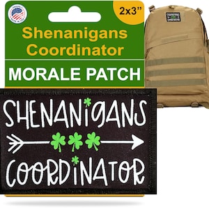 Peut inclure: Un écusson noir avec les mots "SHENANIGANS COORDINATOR" en blanc, une flèche et trois trèfles verts. Un sac à dos beige est en arrière-plan, et une étiquette verte indique "Shenanigans Coordinator Morale Patch". L'écusson mesure 5 x 7.6 cm.