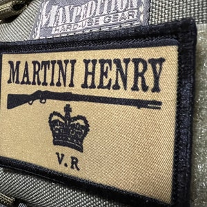 May include: A tan and black patch with the text "MARTINI HENRY" in black lettering. A black silhouette of a rifle with a crown above it and the letters "V.R." below it.