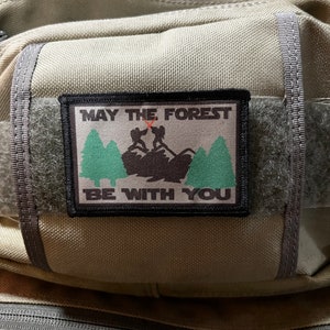 Könnte beinhalten: Ein schwarzer und grüner Patch mit dem Text "Möge der Wald mit dir sein" und einer Silhouette von zwei Personen, die in einem Wald wandern.