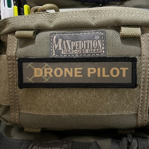 May include: A green tactical bag with a black patch that says "Maxpedition Hard-Use Gear" and a smaller black patch that says "Drone Pilot".