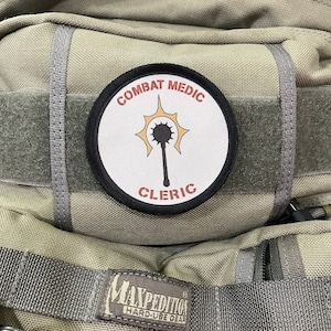 Puede incluir: Una mochila beige con un parche blanco y negro que dice "Combat Medic Cleric" con una estrella roja y una maza. La mochila tiene una correa negra y una etiqueta gris Maxpedition Hard-Use Gear.